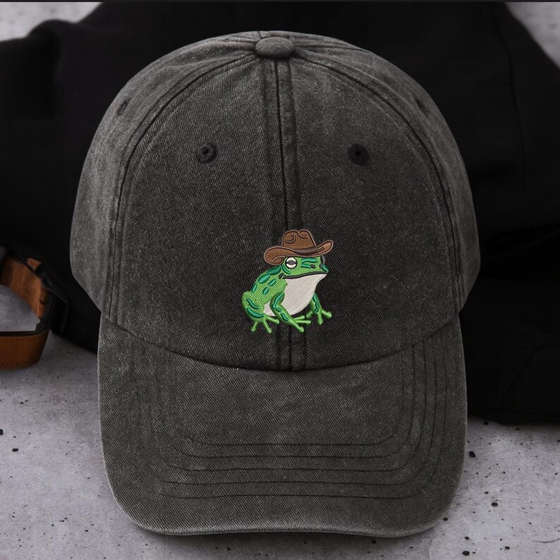 May include: A dark grey baseball cap with a curved peak. The cap has an embroidered green frog wearing a brown cowboy hat. The frog has white detailing and is centered on the front. The cap is made of a textured fabric.