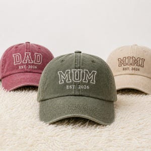Puede incluir: Tres gorras de béisbol en burdeos, verde oliva y beige. Cada gorra tiene texto blanco: "DAD EST. 2026", "MUM EST. 2026" y "MIMI EST. 2026". Las gorras están sobre una superficie blanca texturizada.