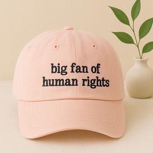 May include: A light pink baseball cap with the phrase "big fan of human rights" embroidered in black. The cap has a curved brim and a rounded top with a small button. The hat is made of a soft fabric.