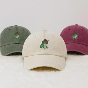May include: Three baseball caps in olive green, cream, and burgundy. Each cap features an embroidered green frog wearing a brown cowboy hat. The caps are made of a soft fabric and have a curved peak.