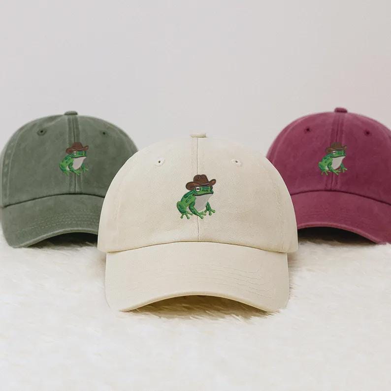 May include: Three baseball caps in olive green, cream, and burgundy. Each cap features an embroidered green frog wearing a brown cowboy hat. The caps are made of a soft fabric and have a curved peak.