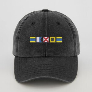 May include: A dark gray baseball cap with a curved brim. The front of the cap features a colorful embroidered design of nautical signal flags. The flags are yellow, blue, white, red, and black. The cap has a classic, casual style.