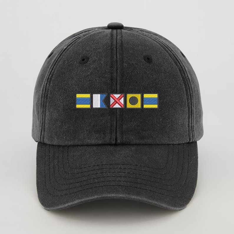 May include: A dark gray baseball cap with a curved brim. The front of the cap features a colorful embroidered design of nautical signal flags. The flags are yellow, blue, white, red, and black. The cap has a classic, casual style.