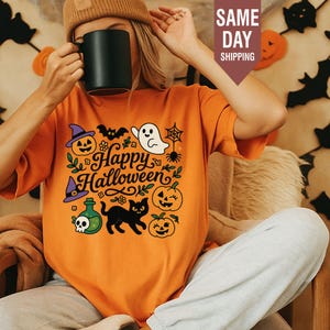 May include: An orange t-shirt featuring a graphic print with the text Happy Halloween, surrounded by illustrations of a jack-o'-lantern, a ghost, a spider web, a black cat, a potion bottle with a skull, and a witch hat in purple and black.