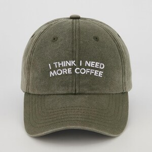 May include: An olive green baseball cap with the embroidered text "I THINK I NEED MORE COFFEE" in white. The cap features a curved peak and a fabric-covered button. The hat is made of a soft, textured material.