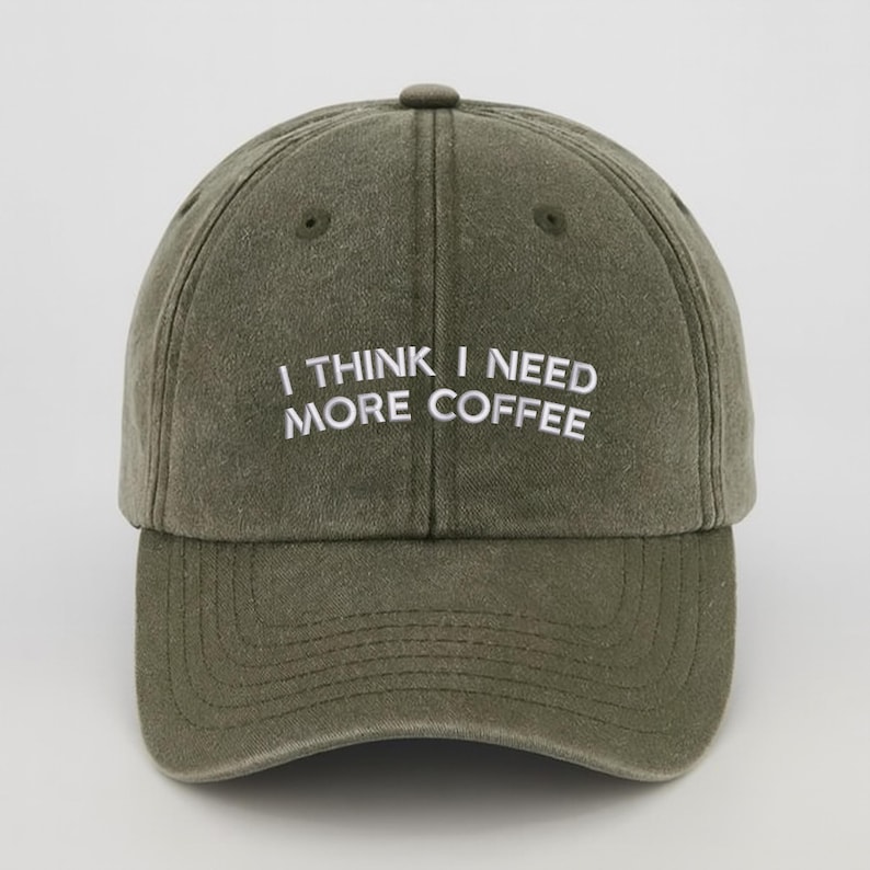 May include: An olive green baseball cap with the embroidered text "I THINK I NEED MORE COFFEE" in white. The cap features a curved peak and a fabric-covered button. The hat is made of a soft, textured material.