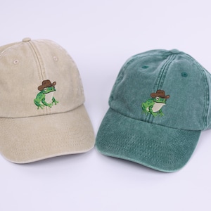 May include: Two baseball caps, one beige and one teal, each embroidered with a green frog wearing a brown cowboy hat. The caps have a curved peak and adjustable strap.