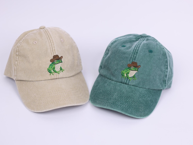 May include: Two baseball caps, one beige and one teal, each embroidered with a green frog wearing a brown cowboy hat. The caps have a curved peak and adjustable strap.