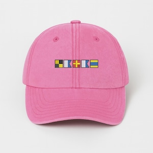 May include: A pink baseball cap with a curved brim. The cap features a rectangular design with colorful nautical signal flags. The flags are in black, yellow, white, blue, and red. The cap is made of a soft material.