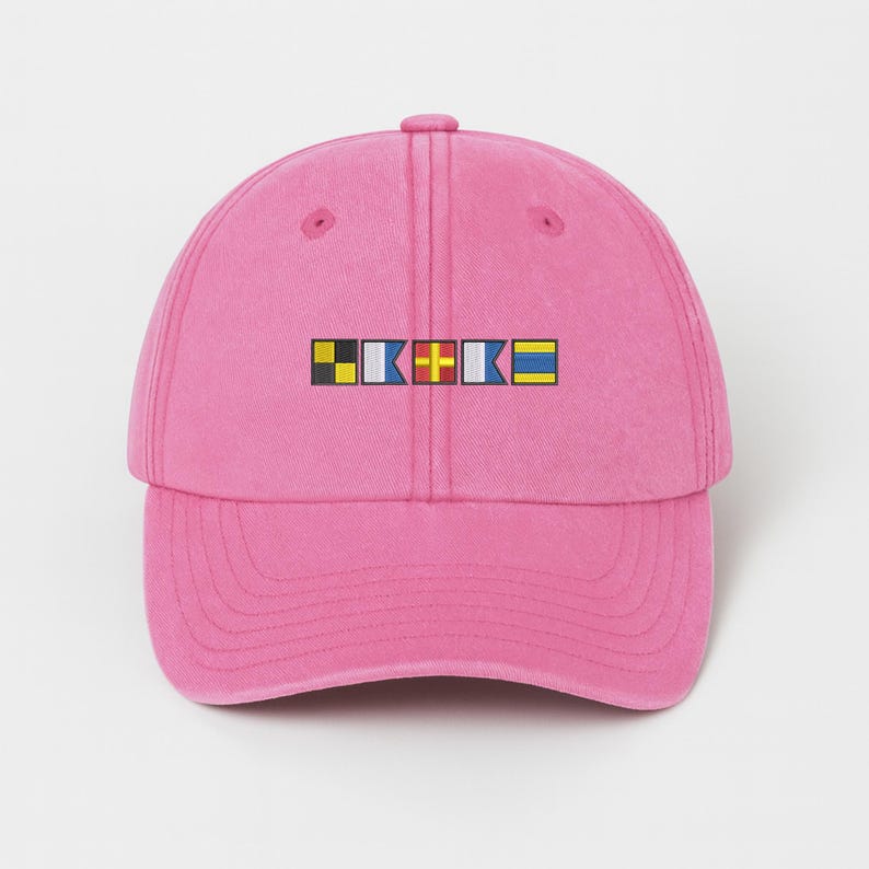May include: A pink baseball cap with a curved brim. The cap features a rectangular design with colorful nautical signal flags. The flags are in black, yellow, white, blue, and red. The cap is made of a soft material.
