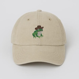May include: A beige baseball cap with an embroidered design of a green frog wearing a brown cowboy hat. The cap has a curved peak and a button on top. The frog design is centered on the front.