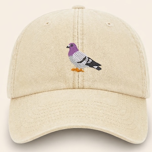 May include: A beige baseball cap with a curved brim. The cap features an embroidered pigeon design in purple, gray, black, and orange. The hat is made of a soft, textured fabric.