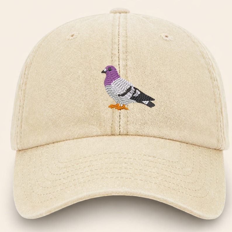 May include: A beige baseball cap with a curved brim. The cap features an embroidered pigeon design in purple, gray, black, and orange. The hat is made of a soft, textured fabric.