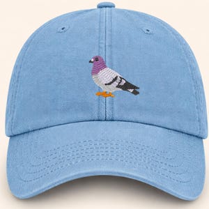 May include: Light blue baseball cap with an embroidered pigeon design. The pigeon has a purple head, gray and white body, black wings, and orange feet. The cap has a curved brim and an adjustable strap.