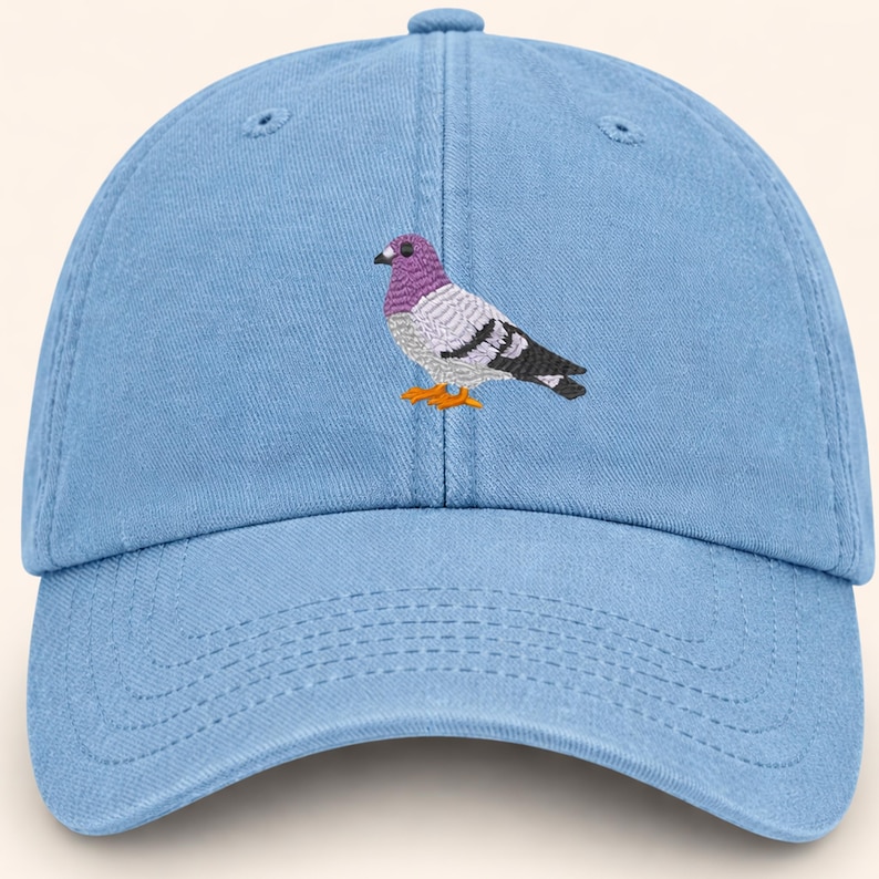 May include: Light blue baseball cap with an embroidered pigeon design. The pigeon has a purple head, gray and white body, black wings, and orange feet. The cap has a curved brim and an adjustable strap.
