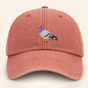 May include: A dusty rose baseball cap with a curved brim. The cap features an embroidered pigeon with purple, gray, black, and orange details. The hat has a six-panel design with a top button and adjustable strap.