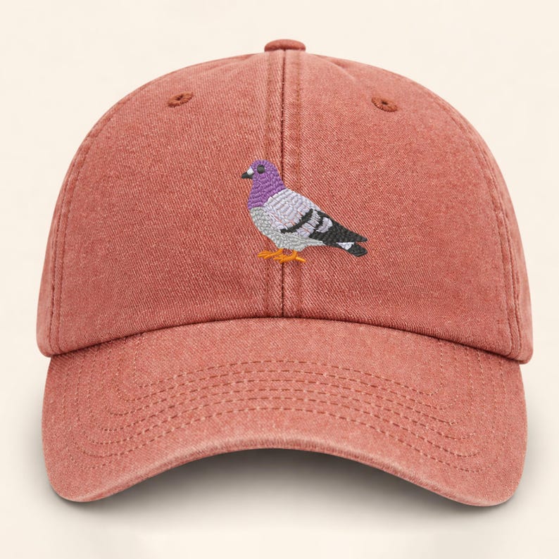 May include: A dusty rose baseball cap with a curved brim. The cap features an embroidered pigeon with purple, gray, black, and orange details. The hat has a six-panel design with a top button and adjustable strap.