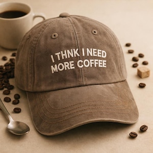 May include: A brown baseball cap with the embroidered text "I THINK I NEED MORE COFFEE" in white. The cap is next to a cup of coffee, coffee beans, and a spoon.