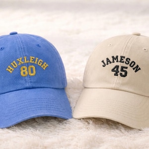 May include: Two baseball caps. One is light blue with "HUXLEIGH 80" embroidered in gold. The other is beige with "JAMESON 45" embroidered in black. Both caps have a curved brim and adjustable strap.