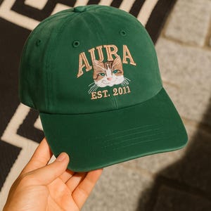 May include: A dark green baseball cap featuring the word "AURA" embroidered in gold above a cat face. The cat has brown and white fur with blue eyes. The text "EST. 2011" is below the cat. The cap is made of a soft material.