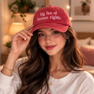 May include: A red baseball cap with the text "big fan of human rights" embroidered in white. The cap is made of a soft material and has a curved brim. The hat is a casual accessory.