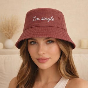 May include: A burgundy bucket hat with the embroidered text "I'm single". The hat is made of a soft fabric and has a classic, casual style. The hat is a solid color and has a wide brim.