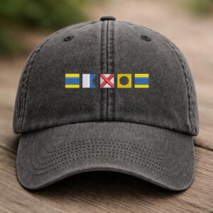 May include: A dark gray baseball cap with embroidered nautical signal flags in yellow, white, red, and black. The cap has a curved brim and a fabric strap closure.