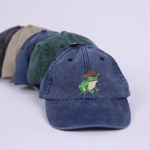 May include: A selection of baseball caps in shades of grey, beige, and blue. The front cap is a faded navy blue, featuring an embroidered green frog wearing a brown cowboy hat. The caps have a worn, vintage appearance.