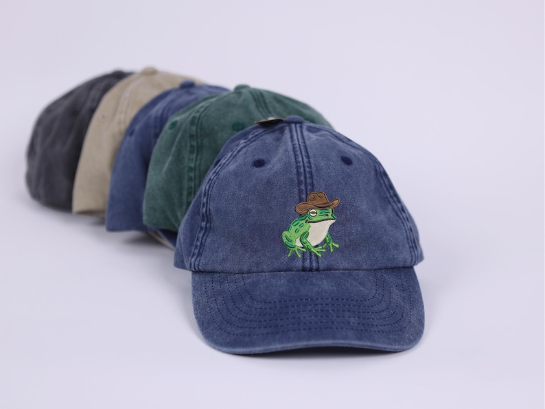 May include: A selection of baseball caps in shades of grey, beige, and blue. The front cap is a faded navy blue, featuring an embroidered green frog wearing a brown cowboy hat. The caps have a worn, vintage appearance.