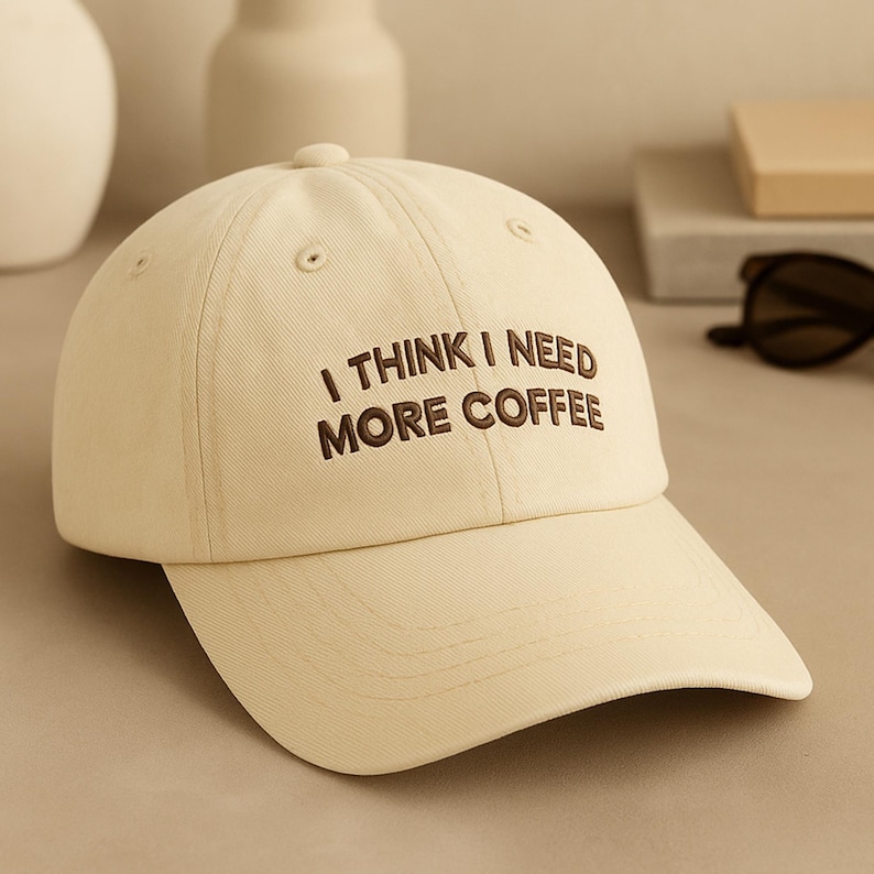 May include: A cream-coloured baseball cap with the embroidered text "I THINK I NEED MORE COFFEE" in brown. The cap has a curved brim and a button on top. The background includes neutral-toned decor.