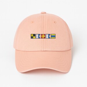May include: A light peach-colored baseball cap with a curved brim. The cap features a small embroidered design of colorful nautical signal flags. The flags are in black, white, blue, red, yellow, and blue. The cap is made of a soft fabric.
