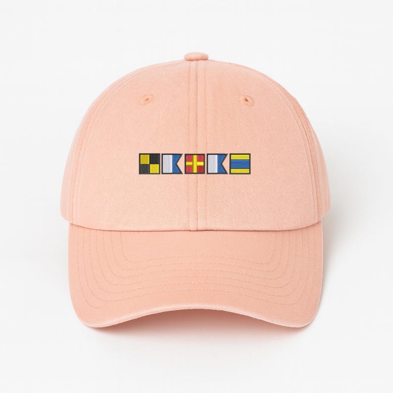 May include: A light peach-colored baseball cap with a curved brim. The cap features a small embroidered design of colorful nautical signal flags. The flags are in black, white, blue, red, yellow, and blue. The cap is made of a soft fabric.