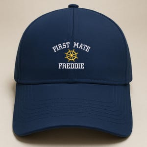 May include: Navy blue baseball cap with the text "FIRST MATE" above a yellow ship's wheel and the name "FREDDIE" in white. The cap has a curved brim and a button on top.