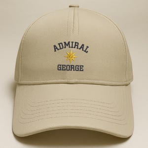 May include: A tan baseball cap with the text "ADMIRAL GEORGE" embroidered in navy blue and gold. The cap has a curved brim and a button on top. The text is arched above a gold sunburst design.
