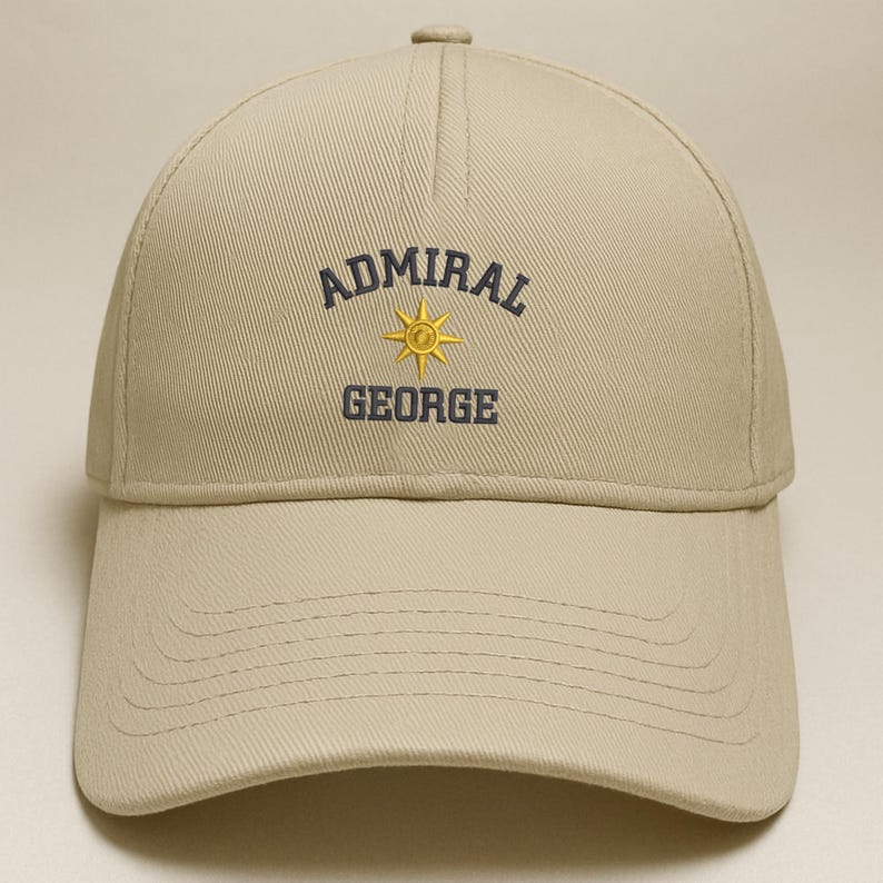 May include: A tan baseball cap with the text "ADMIRAL GEORGE" embroidered in navy blue and gold. The cap has a curved brim and a button on top. The text is arched above a gold sunburst design.