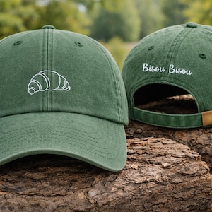 Puede incluir: Gorra de b&eacute;isbol verde con un croissant blanco bordado en la parte delantera. La parte trasera de la gorra tiene el texto "Bisou Bisou" bordado en blanco. La gorra tiene una visera curva y una correa ajustable.