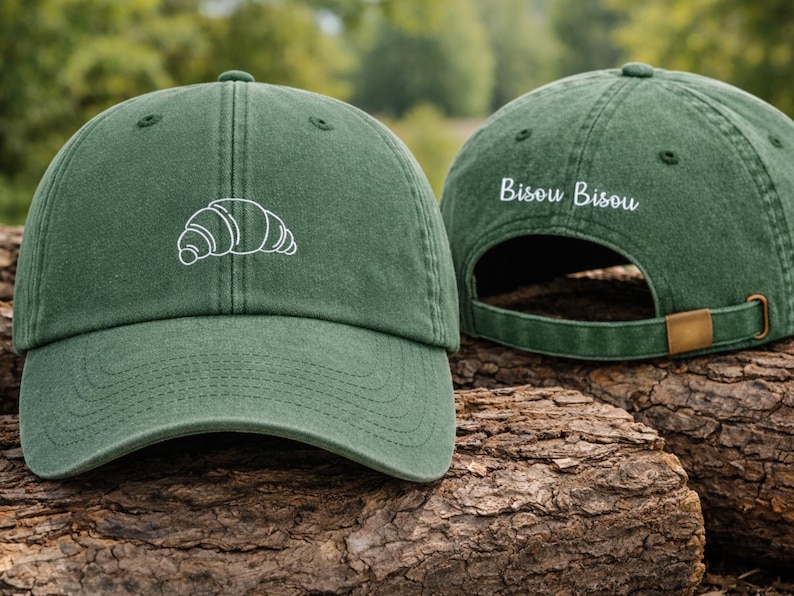 Puede incluir: Gorra de b&eacute;isbol verde con un croissant blanco bordado en la parte delantera. La parte trasera de la gorra tiene el texto "Bisou Bisou" bordado en blanco. La gorra tiene una visera curva y una correa ajustable.