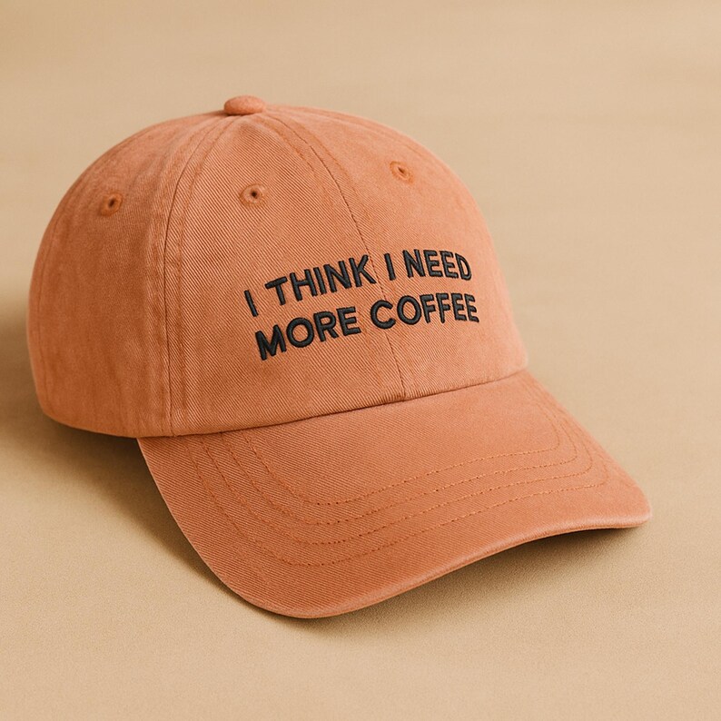 May include: A terracotta-coloured baseball cap featuring the embroidered text "I THINK I NEED MORE COFFEE" in black. The cap has a curved peak and a button on top. The hat is made of a soft, textured material.