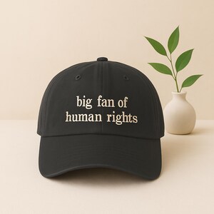 May include: Black baseball cap with the embroidered text "big fan of human rights" in white. The cap is a classic six-panel design with a curved brim and an adjustable strap. A simple and stylish accessory.
