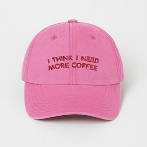 May include: A pink baseball cap with the embroidered text "I THINK I NEED MORE COFFEE" in a darker shade of pink. The cap has a curved brim and a button on top.
