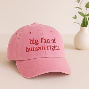 May include: A pink baseball cap with the embroidered text "big fan of human rights" in red. The cap has a curved brim and a button on top. The background includes a white vase with greenery.