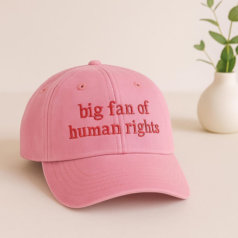 May include: A pink baseball cap with the embroidered text "big fan of human rights" in red. The cap has a curved brim and a button on top. The background includes a white vase with greenery.