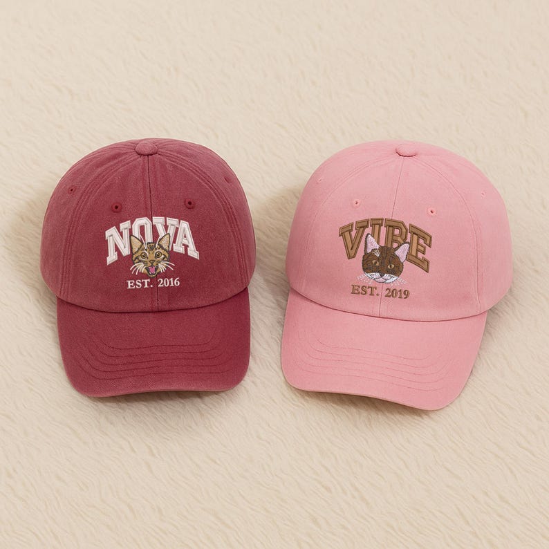 May include: Two baseball caps. One is maroon with "NOVA" and a cat graphic, EST. 2016. The other is pink with "VIBE" and a cat graphic, EST. 2019. Both caps have curved brims and adjustable straps.