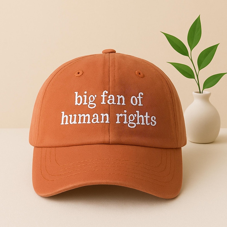 May include: A burnt orange baseball cap with the phrase "big fan of human rights" embroidered in white. The cap has a curved brim and a rounded crown. The background is a neutral beige.