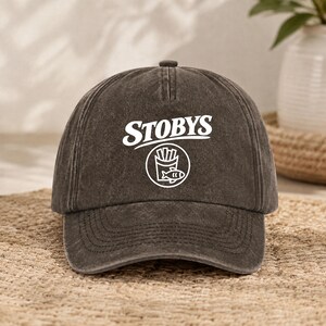 Puede incluir: Una gorra de b&eacute;isbol gris carb&oacute;n con un acabado desgastado. La parte frontal presenta el texto blanco STOBYS sobre un gr&aacute;fico circular que contiene una ilustraci&oacute;n estilizada de un cartucho de patatas fritas y un pescado.