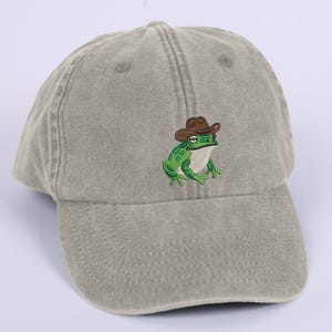 May include: A sage green baseball cap with an embroidered cartoon frog wearing a brown cowboy hat. The frog is green with white detailing. The cap has a curved peak and a classic six-panel design.