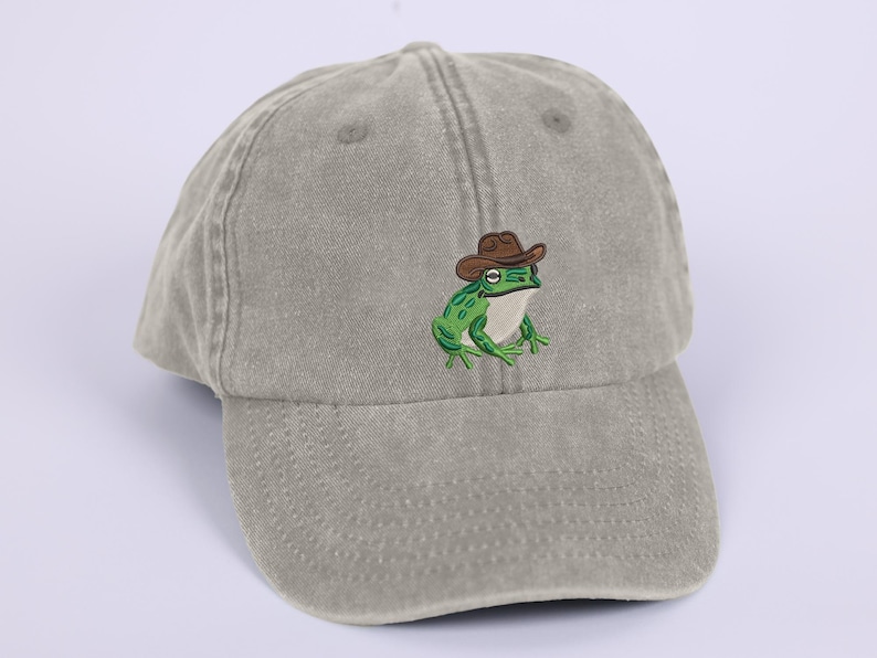May include: A sage green baseball cap with an embroidered cartoon frog wearing a brown cowboy hat. The frog is green with white detailing. The cap has a curved peak and a classic six-panel design.