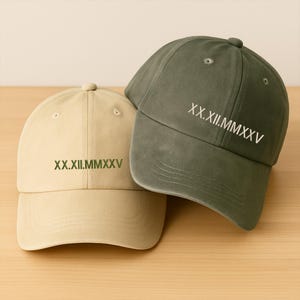 May include: Two baseball caps, one beige and one olive green, are displayed. Both caps have the Roman numeral date "XX.XII.MMXXV" embroidered on the front. The caps are made of a soft fabric and have a curved brim.