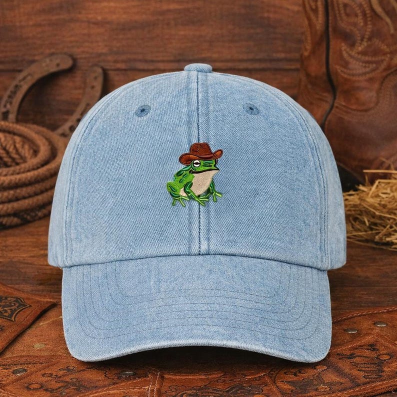 May include: Pale blue denim baseball cap with an embroidered green frog wearing a brown cowboy hat. The cap has a curved peak and adjustable strap. A fun and unique accessory for casual wear.