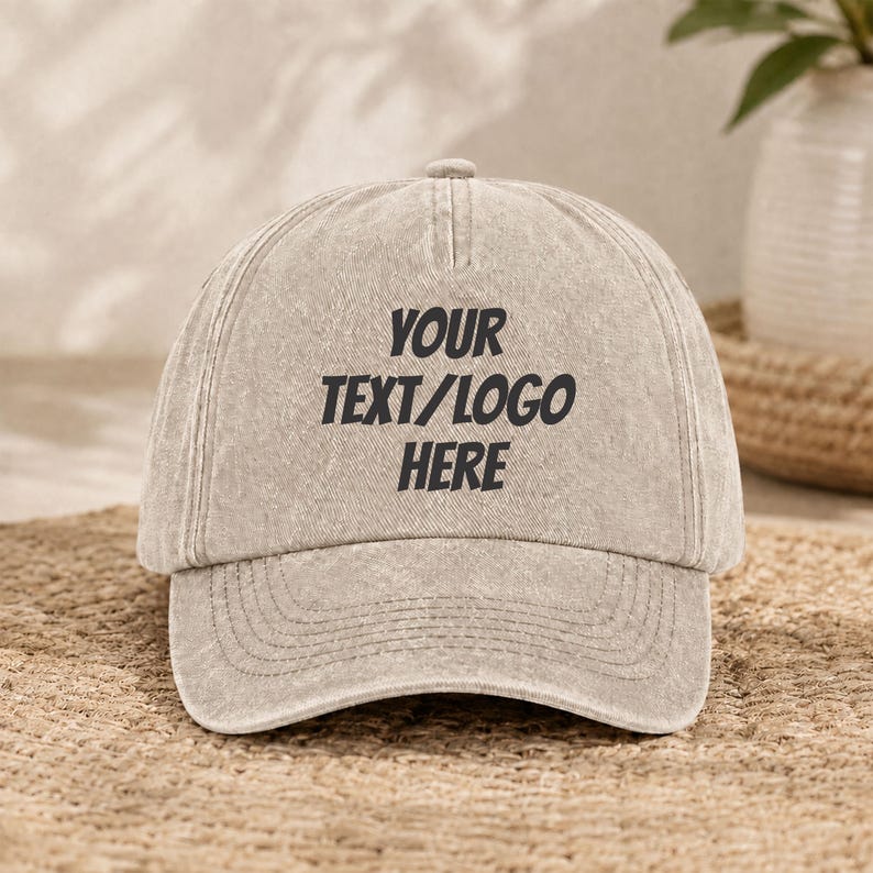 Puede incluir: Una gorra de b&eacute;isbol beige con una textura desgastada y lavada. El panel frontal presenta el texto negro YOUR TEXT/LOGO HERE en una fuente negrita sin remates. La gorra tiene una corona estructurada, una visera curva y costuras visibles.
